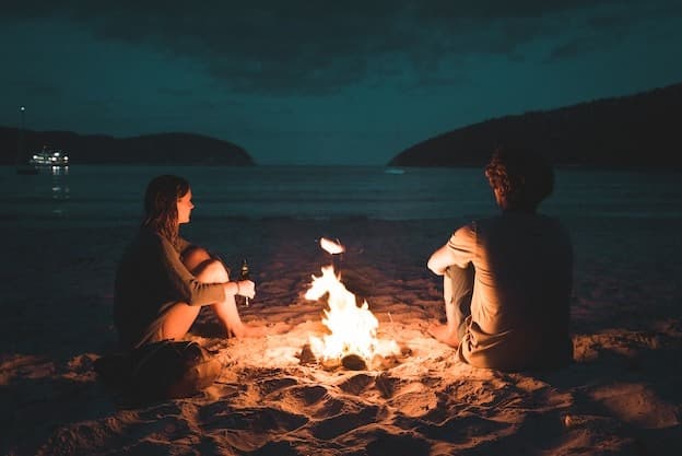 Couple relaxing around warm beach campfire at twilight