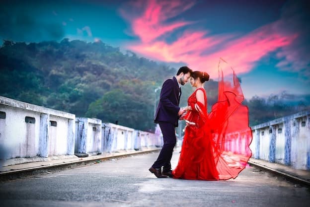 Couple on bridge with flowing crimson gown under sunset sky
